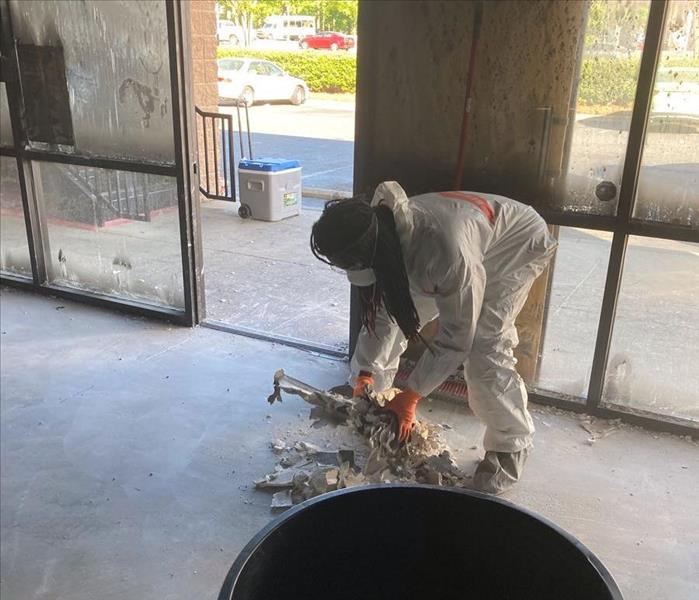 SERVPRO technician in a white tyvek suit, orange gloves and respirator picks up the pile of debris to black trash can.