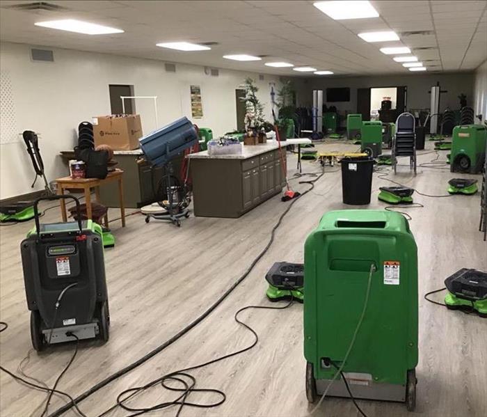 Green SERVPRO air movers and dehumidifiers spread throughout a meeting room with blue chairs, brown/white island.