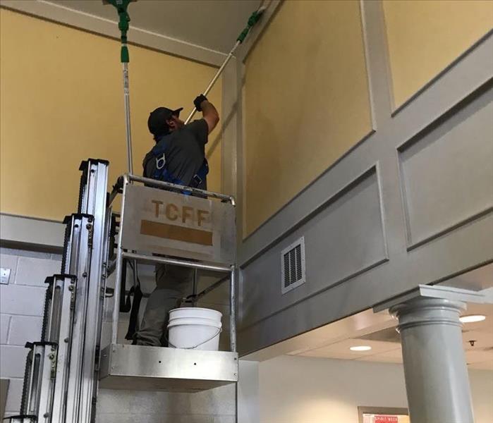 Servpro technician using a vertical mast lift with a green extension pole and chem sponge to clean soot from fire damage. 